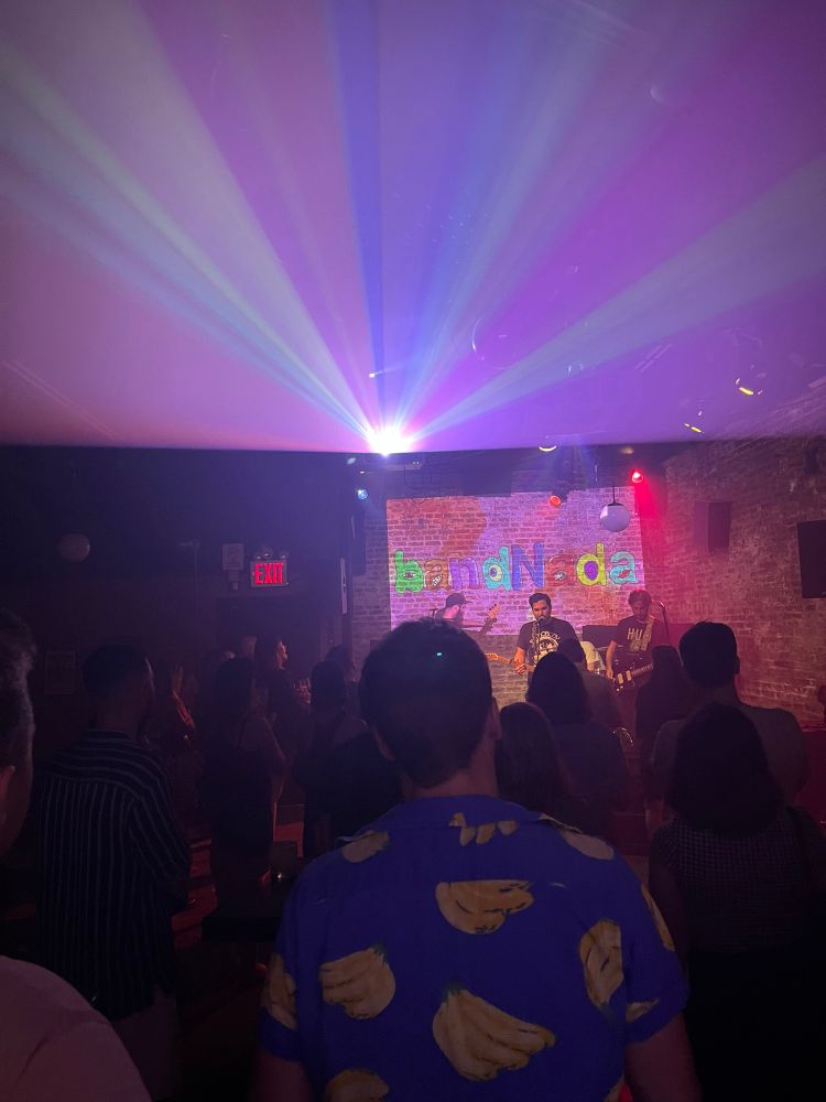 A photo from the crowd at a rock show. The projector overhead creates a fantastic kaleidoscope, and in the foreground a man in a blue shirt with a banana print can be seen from behind 