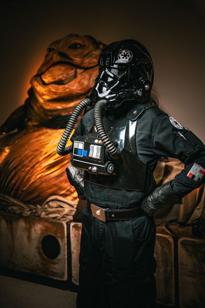 Imperial TIE pilot in glossy black helmet and flight suit stands hands-on-hips in front of Jabba the Hutt display, looking like they’ve landed on the wrong mission.