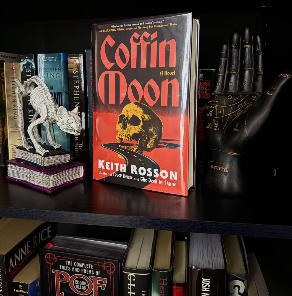 The book Coffin Moon by Keith Rosson sits on a bookshelf facing outward. A skeleton cat statue is next to it and a black palmistry hand. You can see other books behind and below it.
