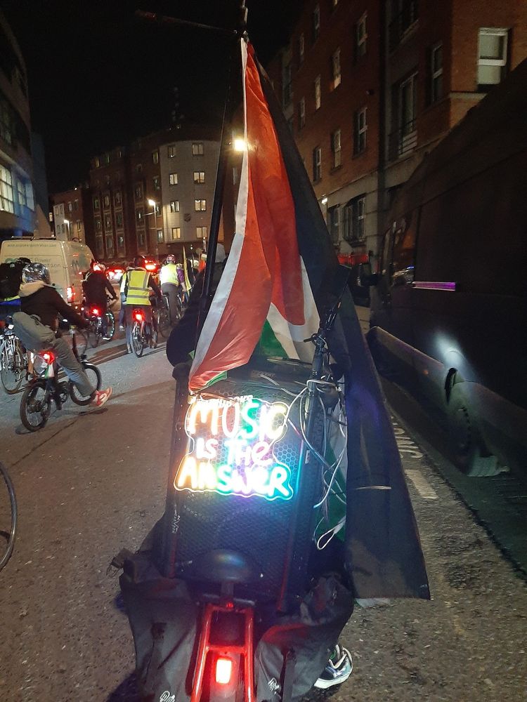 Photo of the back of a bicycle with a large speaker and an illuminated sign saying "MUSIC IS THE ANSWER". There are several other people cycling alongside. There is a Palestinian flag attached to the speaker.