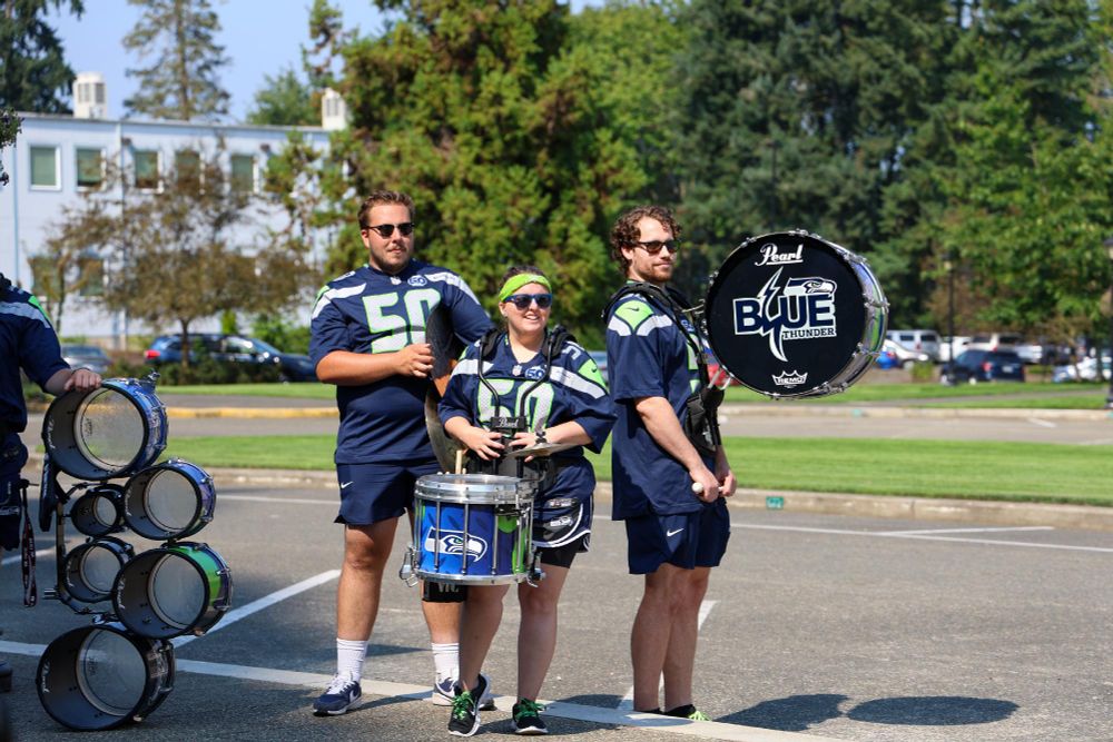 Three individuals in Seattle Seahawks jerseys participate in a drumline outdoors.