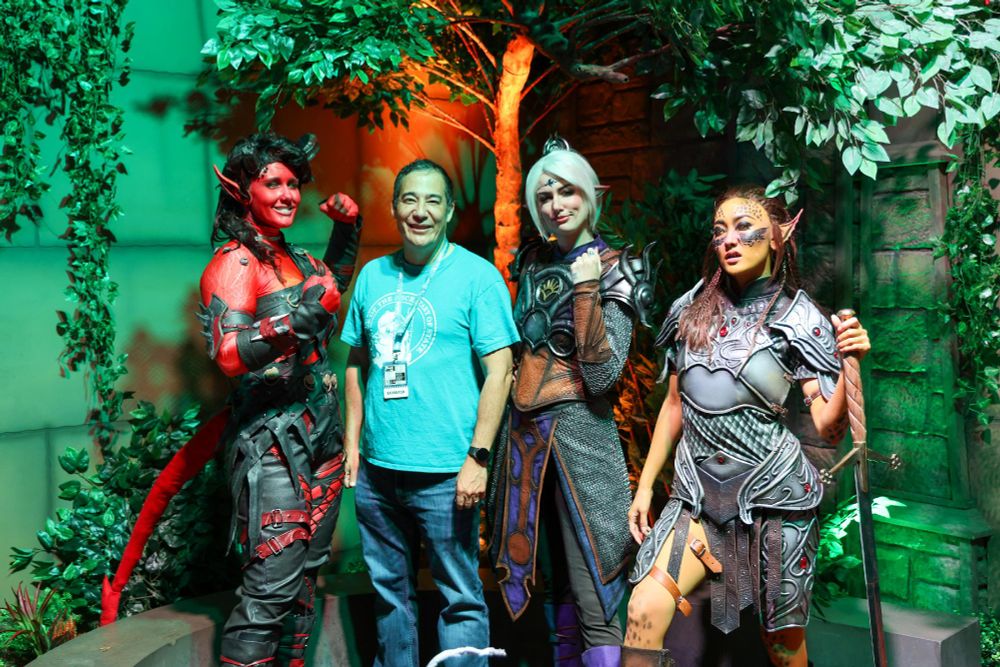 A person stands smiling with three individuals dressed in elaborate fantasy costumes, posing in a set designed to mimic a lush, green forest.