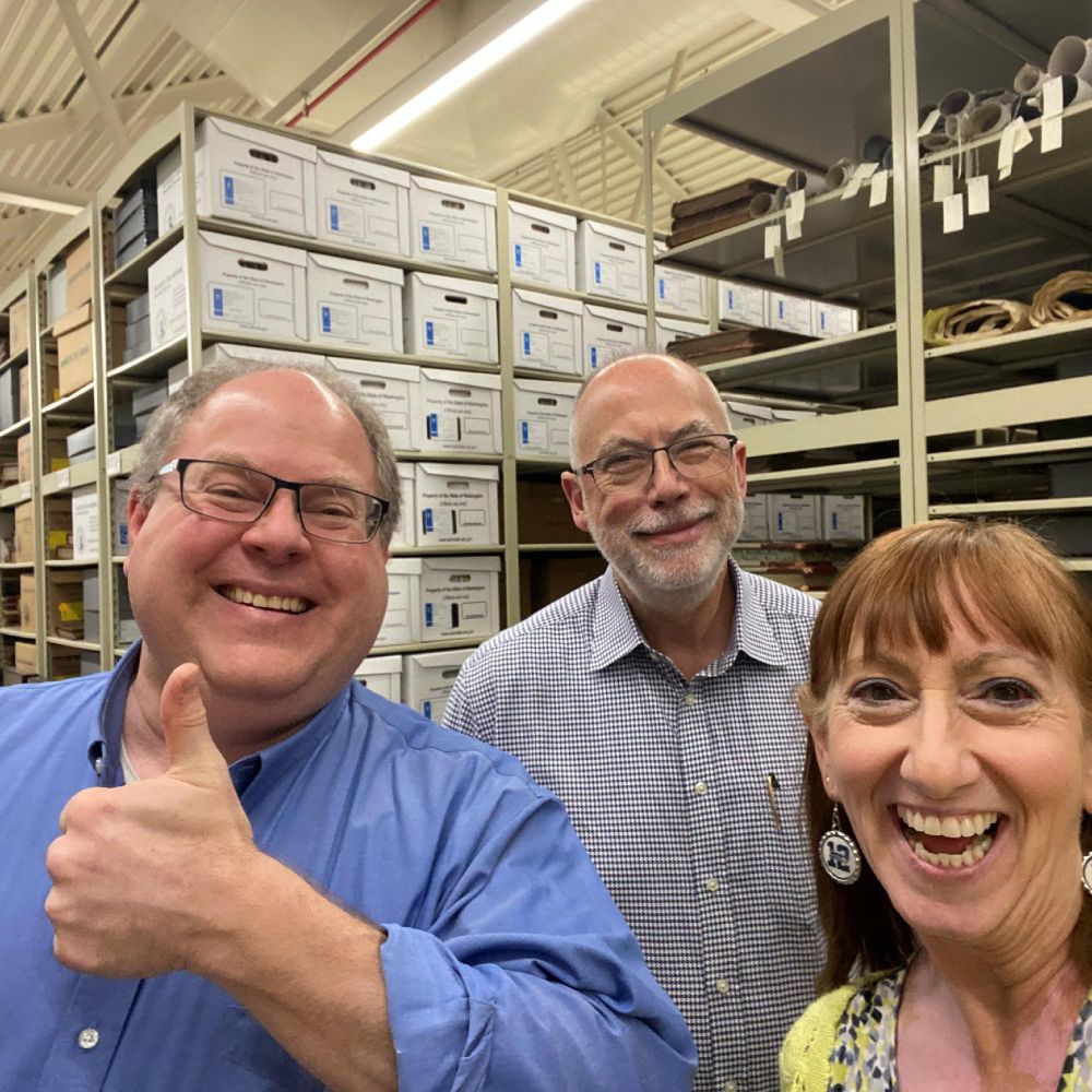 Three smiling individuals posing for a selfie in a storage room filled with boxes and various items. One person is giving a thumbs-up.