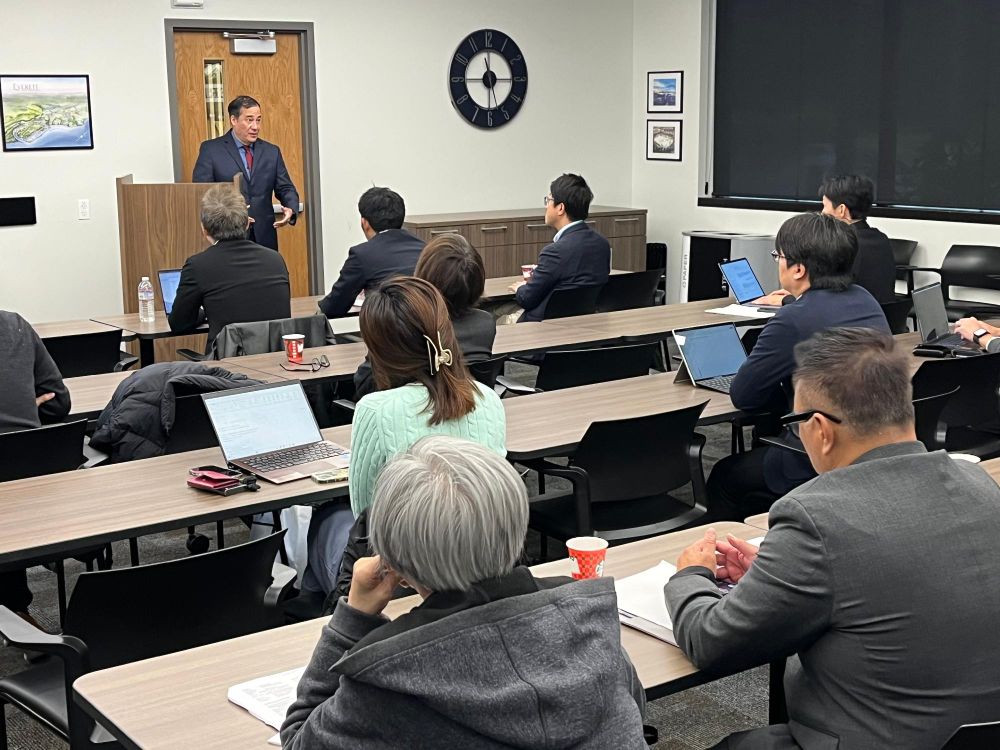 A speaker presenting to a group of attendees in a conference room setting, with laptops open and attentive listeners facing the front where a clock is visible on the wall.