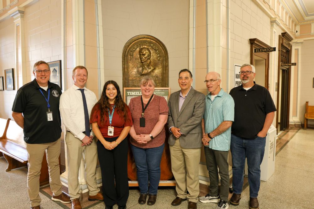  Group of seven individuals standing in a hallway in front of a commemorative plaque at the Lewis County auditor's office.