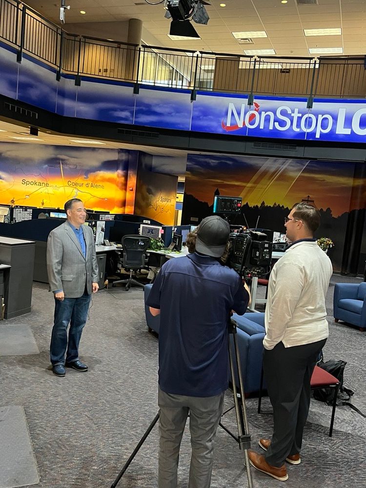 Sec. Hobbs and a group of people in a television studio with monitors displaying "NonStop Local" and city names like Spokane.