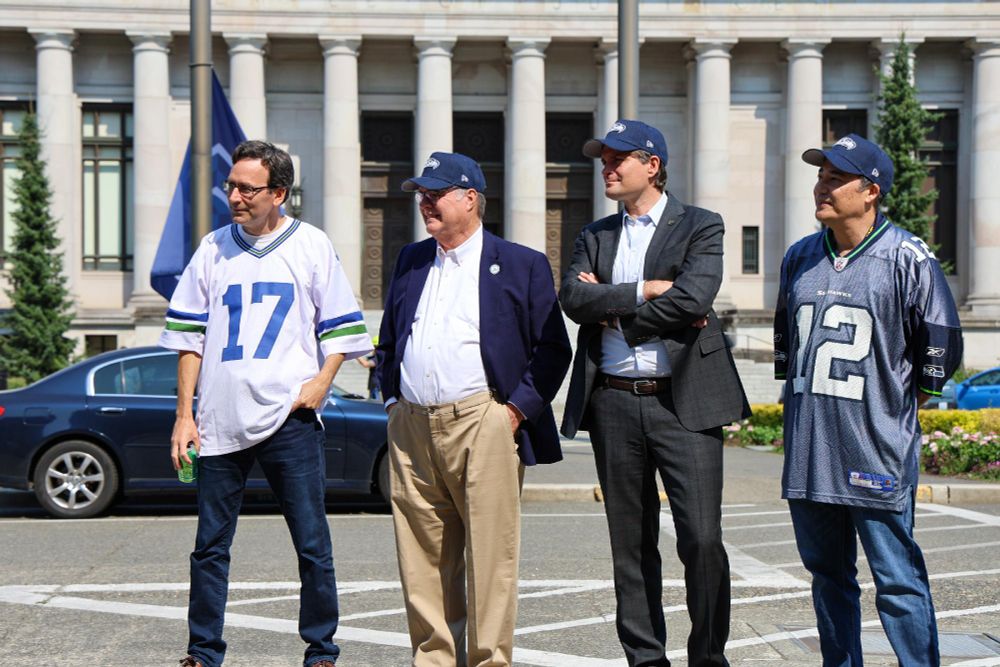Gov. Ferguson, Lt. Gov Heck, Treasurer Mike and Sec. Hobbs wearing Seahawk jerseys stand in front of a government building with flags visible in the background.