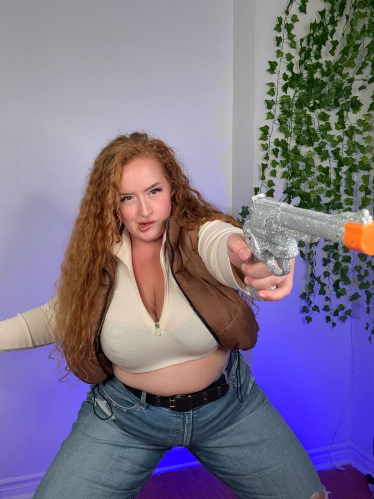Curvy redhead model poses in an action pose as she wears a causal Han Solo cosplay (a brown vest, a cream undershirt and jeans. She is holding a fake sparkly gun and mean mugging as she bites her lip.