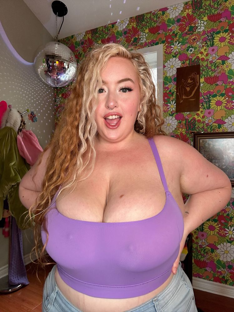 Gwen Adora, a curvy white woman with red curly hair and a blonde money piece, faces the frame waist up. She’s wearing a purple tank top that shows her mid drift, and is sheer to slightly show her nipples through the fabric. She’s looking at the camera with a slightly surprised smile with her hands on her lower back. She’s in a room with floral 60s/70s wallpaper and a disco ball.
