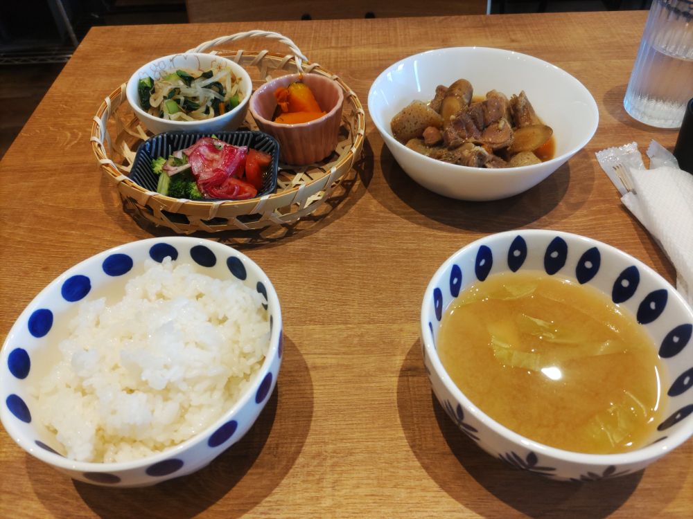 左下から時計回りに、ごはん、汁、肉の煮込み、野菜のおかず盛り合わせが机の上に並んでいる写真