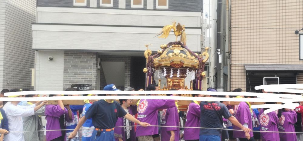 道路を練り歩くお神輿の写真。担ぎ手のほとんどは川崎信金の名前とマークが入った紫色の法被を着ていますが、違う色柄の法被の人も何人か混じっています。