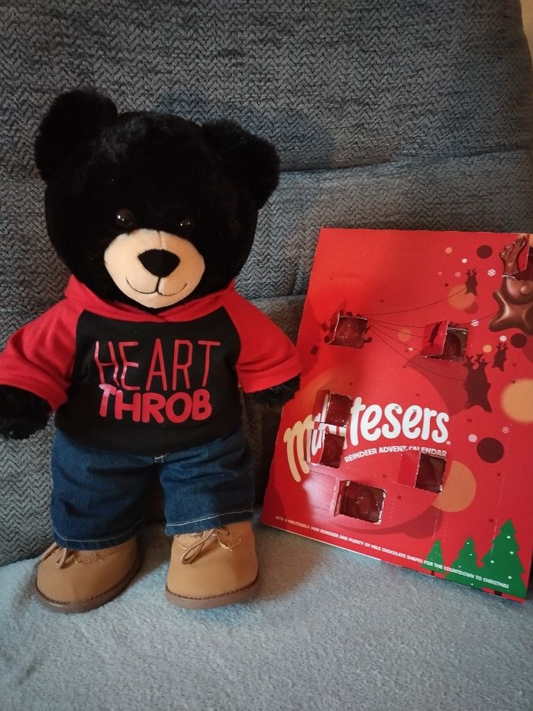 A big black bear called Brian standing next to an advent calendar. He is wearing a red and black top with the words " Heart Throb "  printed on the front, blue jeans and tan boots.