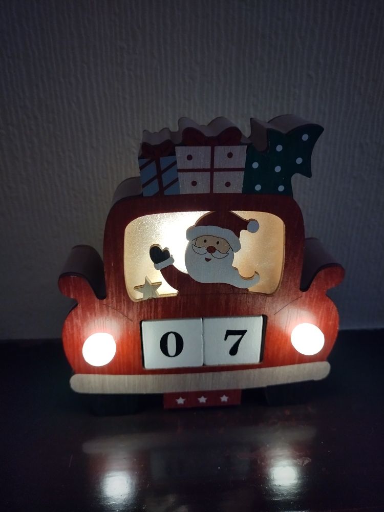 A wooden advent calendar shows Father Christmas driving a red car with presents and a Christmas tree on the roof. The headlights on the car are lit and the number plate is displaying the number seven for the seventh day.