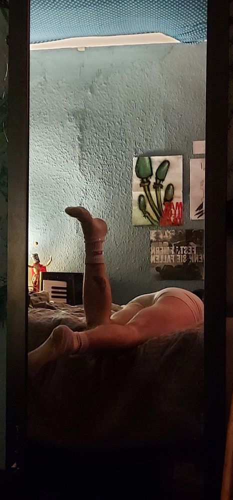 Thw reflection in a mirror shows the slimly muscular legs of a white guy in white boxers and socks laying on a bed 