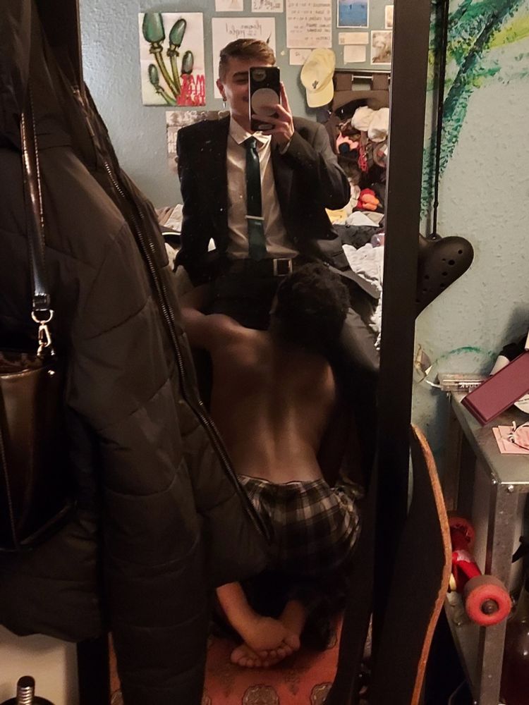 A mirror selfie, of a white guy in a suit sitting down, he has a green tie and his face is covered by the phone. In his lap is a shirtless black guy with his head in his crotch, back arched, booty popping, cute toes out.