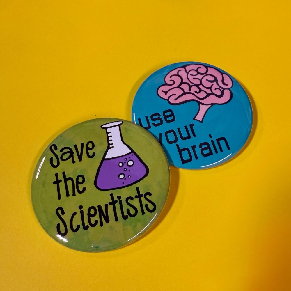 Two pinback buttons on a yellow background. One says save the scientists with a beaker and the other says use your brain with a drawing of a brain. 