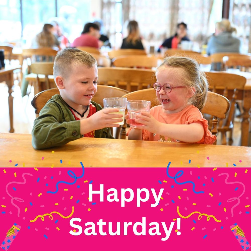 Photo of two young children sitting at table holding their glasses of juice up to 'cheers' and smiling. White Text on bright pink background below reads 'Happy Saturday!', with confetti popper graphics bursting in each bottom corner towards the centre