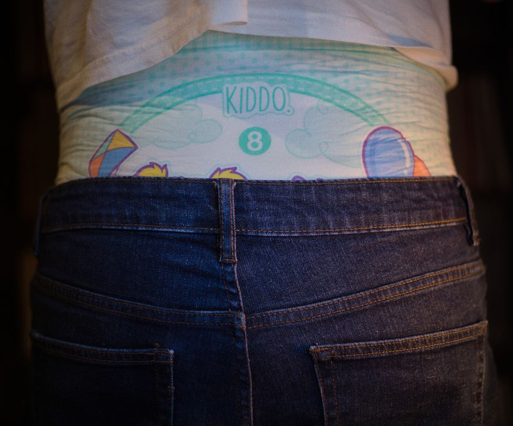 The jeans pulled lower, below the hips. A vibrant print on the adult diaper is now partly visible.