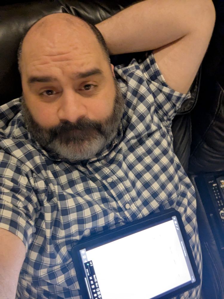 Photo of me, bearded middle aged bald guy, with my arm behind my head, recloned in a blue and white gingham patterned short sleeve button up, with a tablet on my stomach.