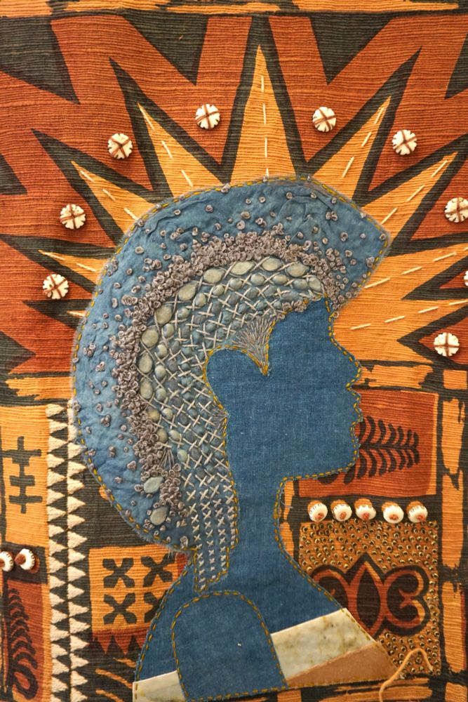 Background features Orange dyed textured fabric with various symbols and patterns in dark blue, with off white embroidery and beads. In the foreground there is a figure made of indigo dyed fabric, with seeds from various plants embroidery into its hair. Half of the hair is still unbraided and in an Afro, and the braiding of the seeds is in process. 