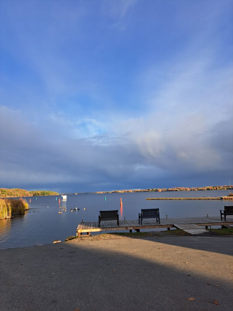 A band of sunlight remains as shadows are cast with the sunset. Park benches face away from the camera, out across the lake. It is the Golden Hour, with a  golden haze everywhere. The sky is blue with heavy cloud coverage. 