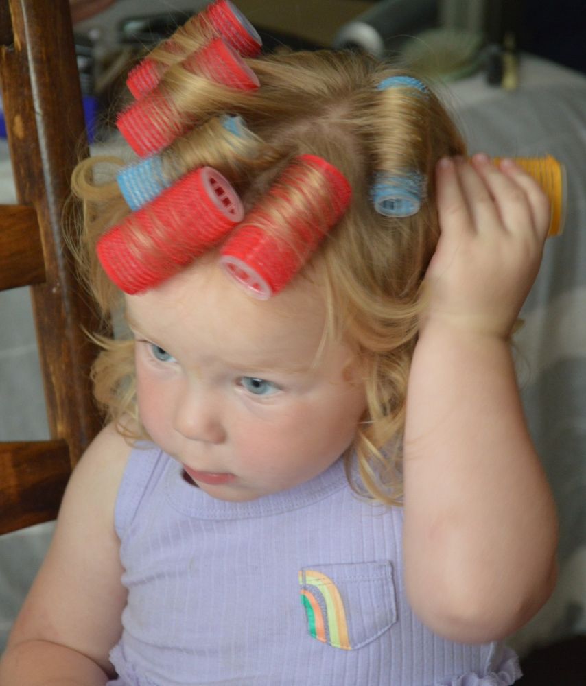 A little girl with strawberry blonde hair, places a yellow velcro curler in her hair. It joins the pink and blue ones. Her dress is light mauve.