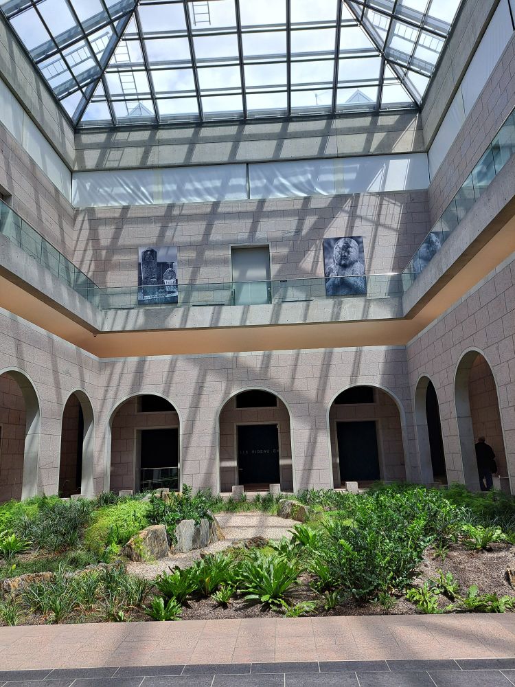 The skylight overhead casts a matrix of shadows across the atrium. The mezzanine, with its glass railing, invite our gaze skywards, with an unobstructed view. A path with gardens filled with foliage on either side of it, completes the frame. 