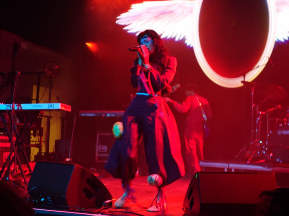 Magdalena Bay performing on a deep red stage with angel wings in the background. Mica Tenenbaum holds the mic with two hands as she sings in a red costume with planets dotted on the costume.