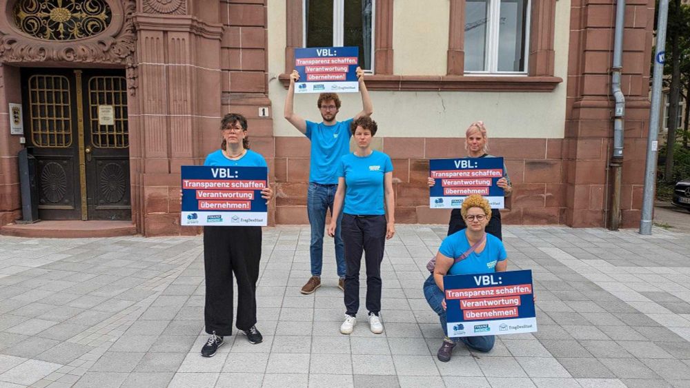 Fünf Personen stehen und knien vor einem historischen Gebäude mit rotem Sandstein. Sie tragen blaue T-Shirts mit dem Logo von FragDenStaat und halten Schilder mit der Aufschrift: „VBL: Transparenz schaffen, Verantwortung übernehmen!“