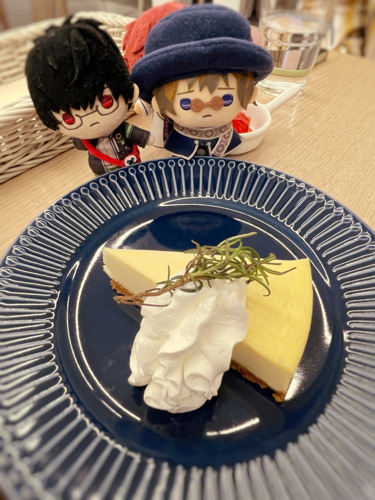 コリーカフェの魔法使いの約束コラボメニュー、ニューヨークチーズケーキをみつめているレノックスさんとファウストさんのぬいぐるみ（を背後で支えているミスラさんのぬいぐるみ）