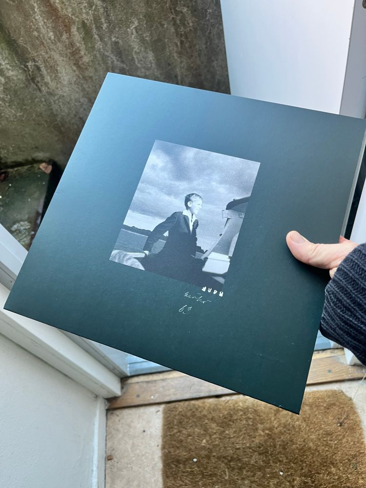 Hand holding an lp sleeve with a black and white photo of a young boy on a boat in the centre of a dark background 