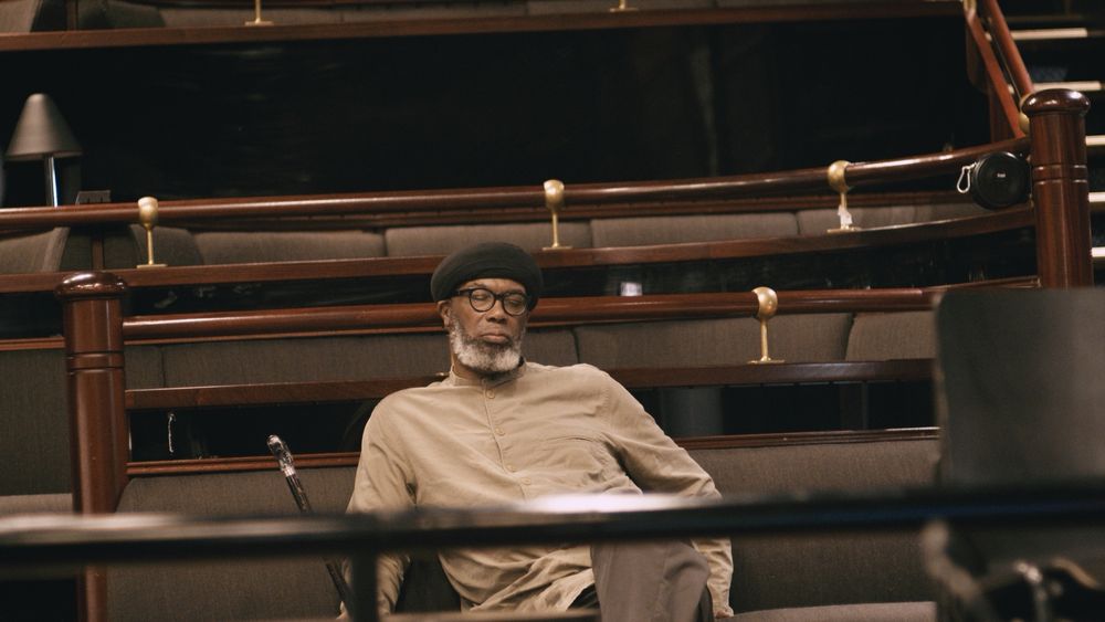 Pat Thomas sits alone amongst rows of empty seating with fine polished timber railings at Victoria, the national jazz theatre in Oslo. He wears a tunic and holds a walking cane in his right hand.