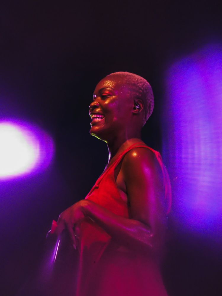 A cantora Pongo sorri amplamente. A iluminação é roxa e rosa, criando um contraste com seu vestido vermelho. Seu rosto brilha com o suor da performance, transmitindo alegria e intensidade no palco.