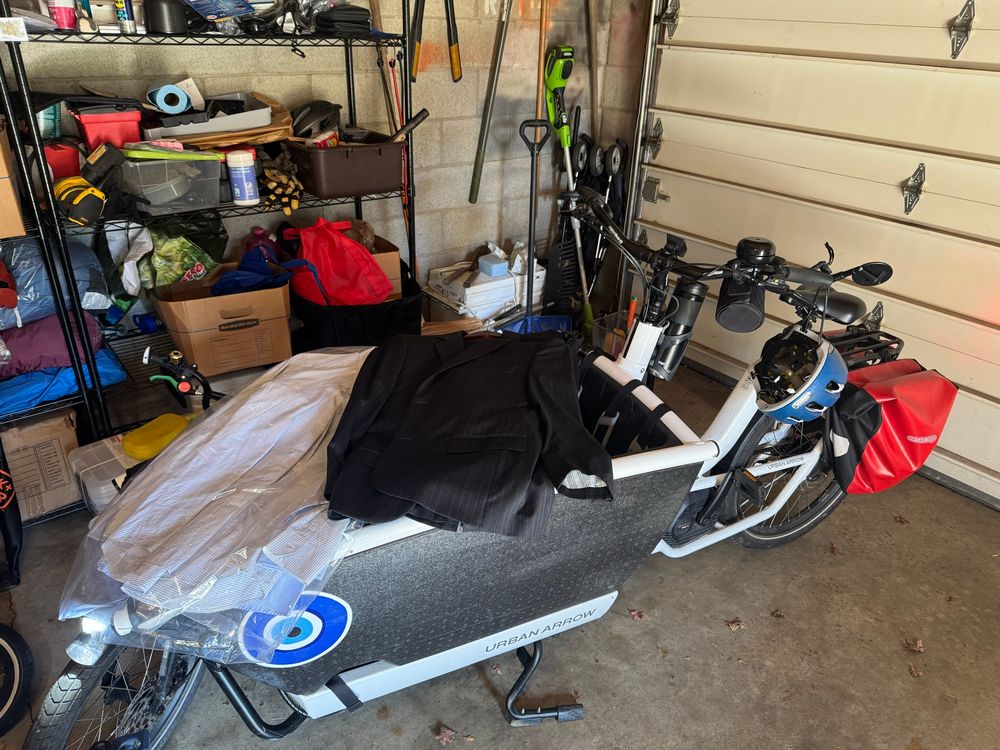 An urban arrow bakfiets parked in a garage with a suit and some dress shirts laid out on the tonneau cover. 