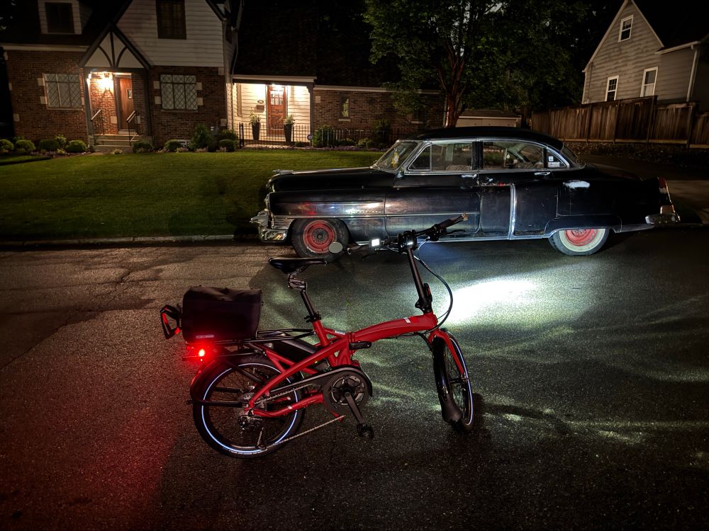A 1930s or 1940s Cadillac parked on the street at night, next to a Tern Vektron e-bike with its lights lit