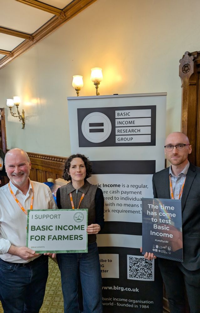 Left to right: Simon Duffy, Jo Poulton, and Elliott Johnson stand in a committee room at the Palace of Westminster, holding posters. One reads 'I Support Basic Income for Farmers,' and the other says 'The Time Has Come to Test Basic Income.'