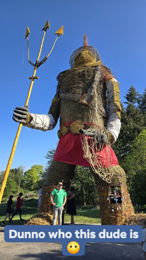 A towering sculpture made from woven natural materials stands against a clear blue sky. The figure resembles a giant warrior or deity, outfitted with a red skirt, a golden belt, a net draped over one arm, and a tall yellow trident in hand. Its head is topped with a red plume and features circular eye cutouts. Several people, including a man in a green shirt standing beneath the figure, give a sense of the sculpture’s immense scale. The setting is a grassy park area with trees and scattered visitors. Overlaid text humorously reads, “Dunno who this dude is 😶.”