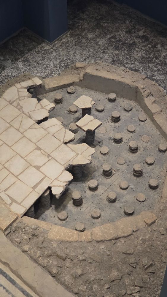Circular hypocaust system on display at the Zeugma Mosaic Museum, showing a partially reconstructed Roman underfloor heating structure. The floor tiles are supported by evenly spaced stacks of round ceramic discs (pilae), revealing the open space beneath where hot air would circulate to warm the room above. The surrounding area includes excavated earth and remnants of the original flooring.

