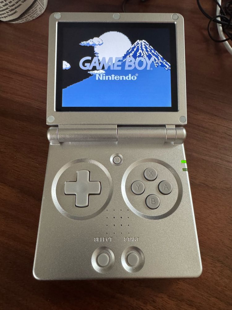 A portable video game system looking like a GBA SP in silver color. 
The four face buttons give away that it’s not a GBA SP (it’s an Anbernic RG35XXSP).

It shows a Nintendo Game Boy splash screen with a pixel-art mount Fuji in shades of blue.