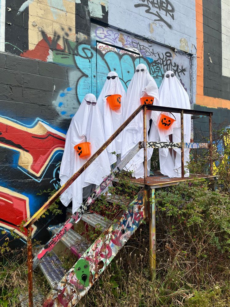Four ghostly figures (people in white sheets with sunglasses) holding pumpkin buckets on a set of steel stairs in front of a colourful wall of graffiti