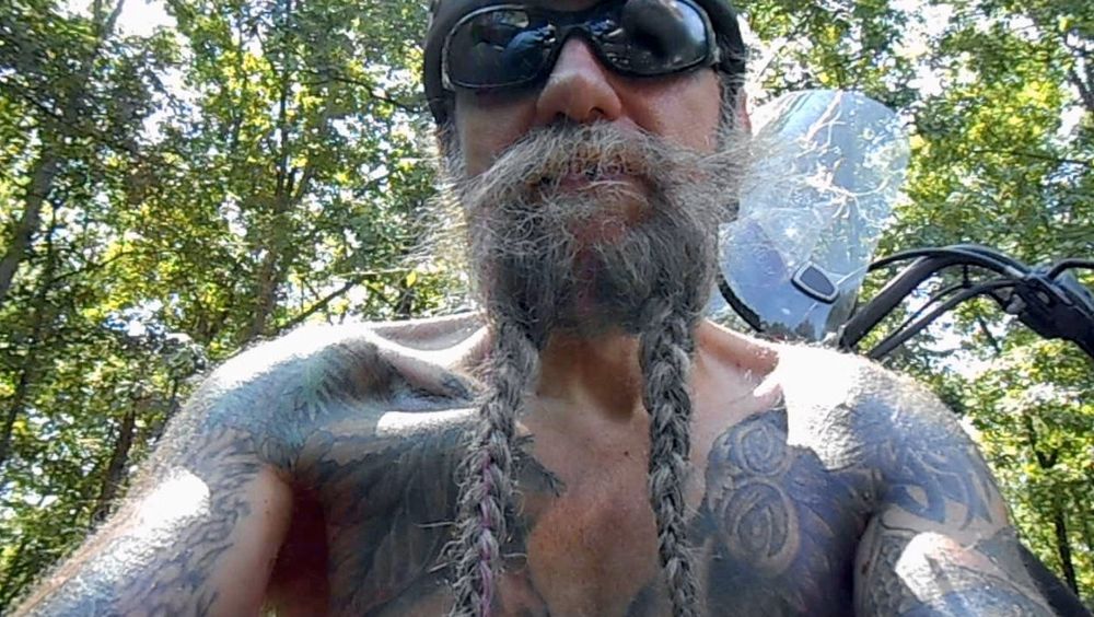 Close up view looking up at a tattoo'd biker. His upper chest arm and face dominate the image. His long beard is divided in two and hangs in a pair of thick ropes from his chin. His wrap around dark glasses vaguely reflect the bike he's working on. 