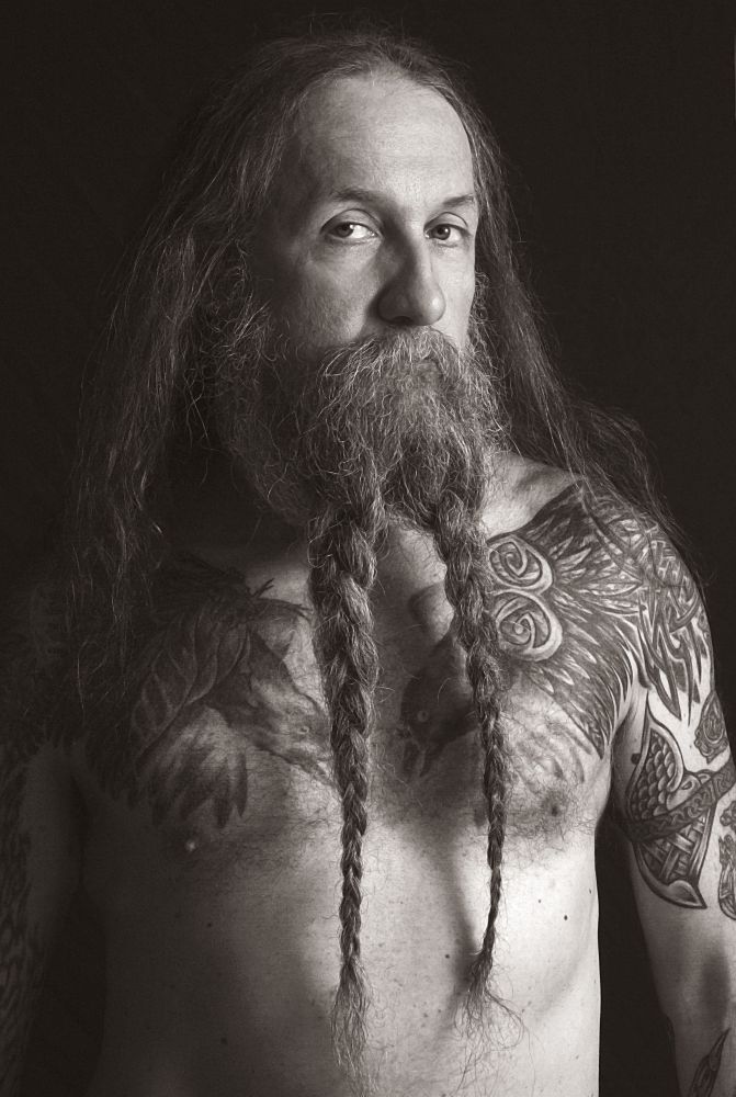 A dramatic photo of a shirtless man with many tattoos. His long beard is in two long braids. His long hair, parted in the middle, hangs below his shoulders. The most notable thing are his eyes. While his head is turned slightly to his left, his eyes stare to his right as if seeing something to left of the camera.