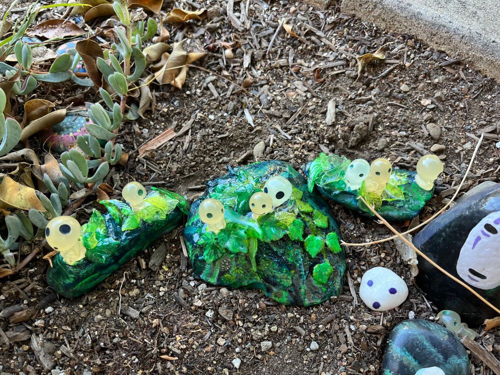 Three large rocks of various irregular shapes  in a garden that are painted to look like forested scenes in deep shades of green, brown, and blue, with three dimensional leaves on top. Sitting atop each of them are two to three plastic figurines of kodama forest spirits from the Ghibli film Princess Mononoke.