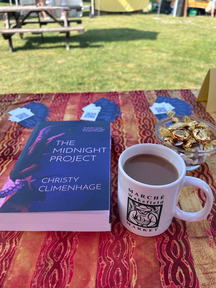 A book with pink tentacles on the cover entitled The Midnight Project by Christy Climenhage sits on a table beside a mug of coffee with “Marché Wakefield Market” and a design of a farmer on it. A bowl of candies and fans of business cards are behind  them.