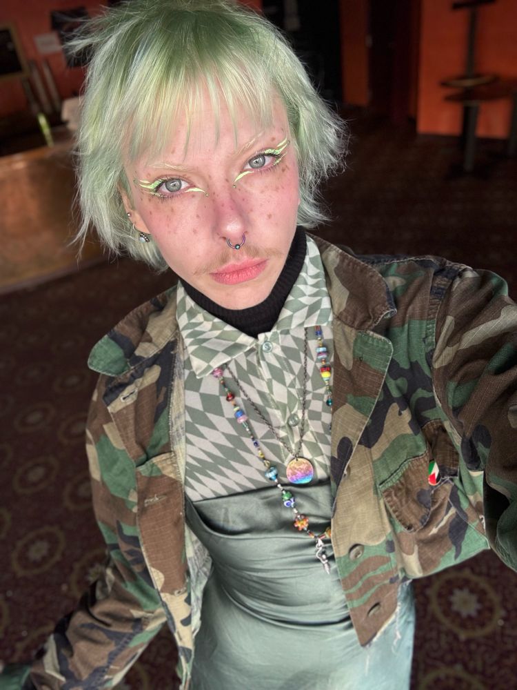 a selfie of a trans masc non-binary individual, with minty green colored hair in a pixie cut, and matching green makeup, and outfit. the outfit is a sage green colored satin dress layered over a duo toned green checkered mesh button up collared dress, and with a cropped camo jacket on top, accessorized with a Palestine flag pin, and layered necklaces.