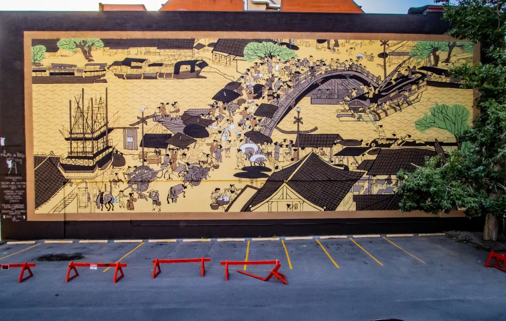 A photography of a massive mural beside a parking lot. The mural is a reimagined version of the scroll Along the River During the Qingming Festival. The mural depicts the Song dynasty in China, stylistically the characters are derpy and whimsical, and the main focus of the mural is a bridge bustling with activity. Vendors sell their wares, government officials are in standoffs, a boat is trying to navigate underneath the bridge, but is close to hitting it. Around the bridge is a river and the edge of a city.
