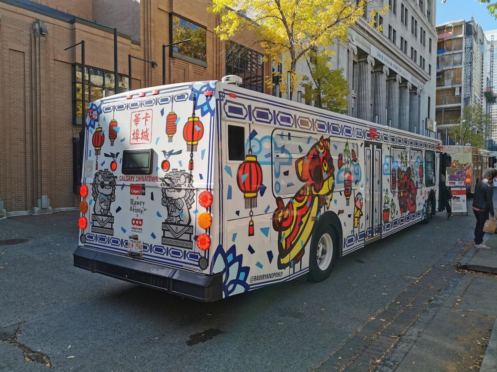 The image depicted is a courtyard with a bus parked in the centre and people milling about the area. The bus has two sides visible, the back and the right side with the door. A tree with fall leaves is in the background. 

On the long side a lion dance with anthropomorphic derpy cabbages, drippy clouds, lanterns, and gongs. The Lions are derpy. Everything is derpy.

On the back there are three logos City of Calgary X Rawry & Pohly X Calgary Arts Development.

Confetti is everywhere. The back has stone lions and magpies. There are patterns all around the bus, similar to Chinese Porcelain patterns found on plates, cups, and bowls that would be found frequently in Chinatown.