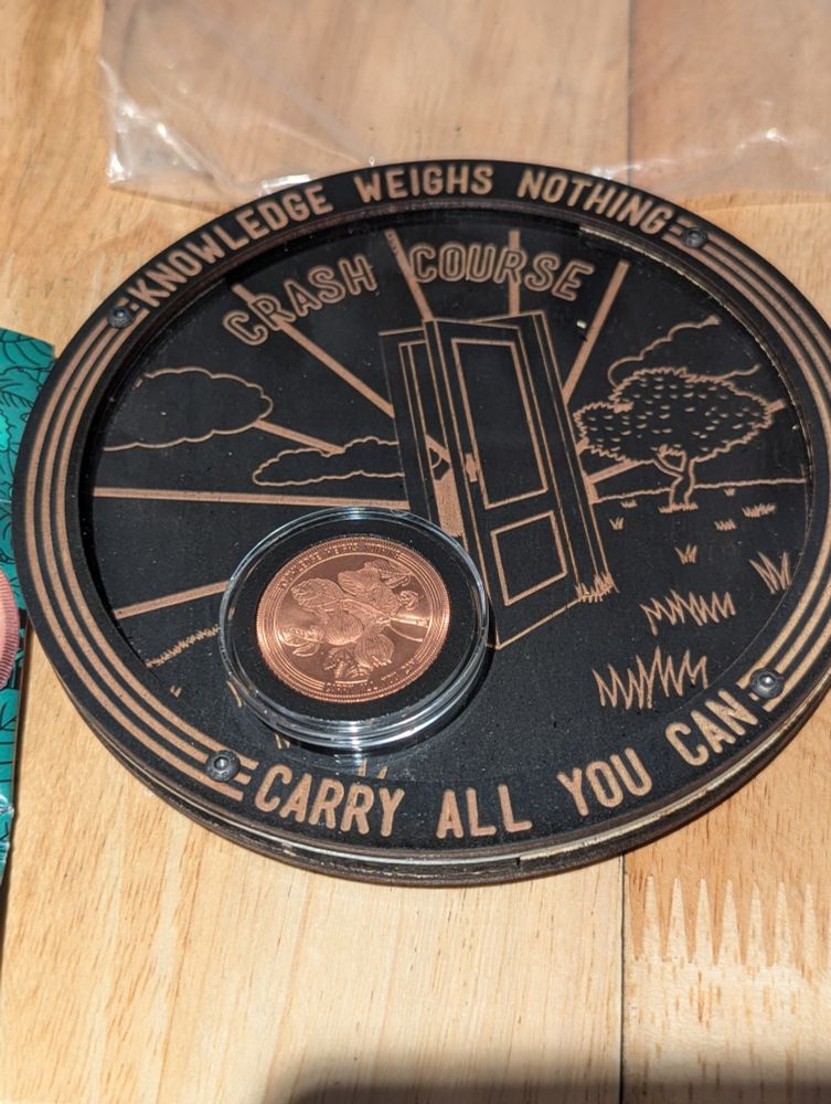 Photo of 2024 Crash Course Coin enclosed in its plastic case. laying on top of the new Crash Course Coin bank. Bank has the Crash Course logo of an opening door in a field, and reads "Knowledge weighs nothing... carry all you can". 