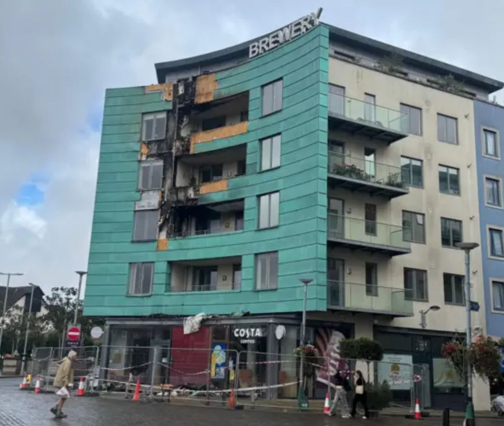 An image of a block of purpose built flats, five upper storeys. The frontage is green due it being constructed using copper wrapped around timber as cladding. Upper storeys 2, 3 and 4 have areas around their balconies that have been removed and exposed, it happened during and after a fire, to ensure it was properly extinguished after it had spread through the timber in the cladding.