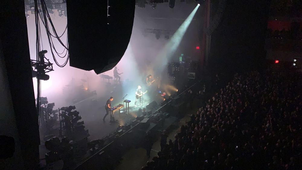 Nine Inch Nails on stage at the hard rock in Las Vegas, taken from above (2nd floor table vip seating). Trent Reznor has a spotlight on him, while rest of crowd and band is mostly dark. Band is dressed in all black, and smoke machine and strobe light effects cover the stage.